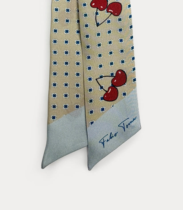 Dettaglio ravvicinato del nastro twilly che mette in risalto il rosso brillante delle ciliegie stampate, la precisione del micro-motivo geometrico blu sullo sfondo beige e la fine trama del tessuto.